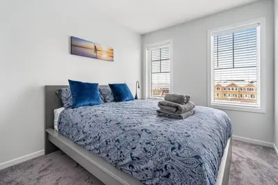a bedroom with a bed with a blue comforter and two windows at 4 Bedroom Town Home with Garage Parking, Fast WiFi, Smart TV with Netflix, Fully Equipped Kitchen, Sleeps 10, Ideal for Families in Airdrie