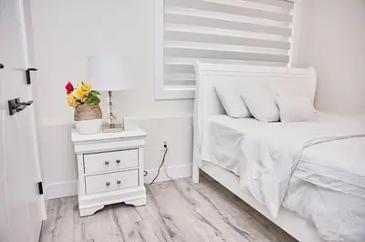 a white bedroom with a bed and a night stand with a lamp at Brand-new three bedroom guest suite in the desirable neighbourhood in Prince George