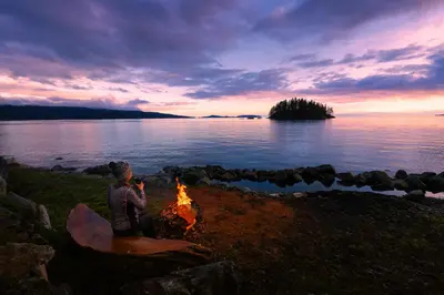 a woman sitting around a campfire on the shore of a lake at Beaver Beach Vacation Home in Madeira Park