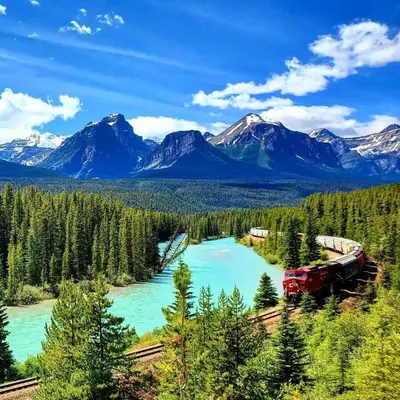 a train on tracks next to a river with mountains at Newly Built 2BR Mountain Escape in DT Canmore! in Canmore