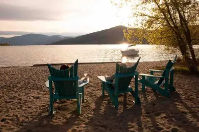 a woman sitting in chairs on a beach with a boat at Top Floor on Okanagan Lake with AC & Heated Pool in Vernon